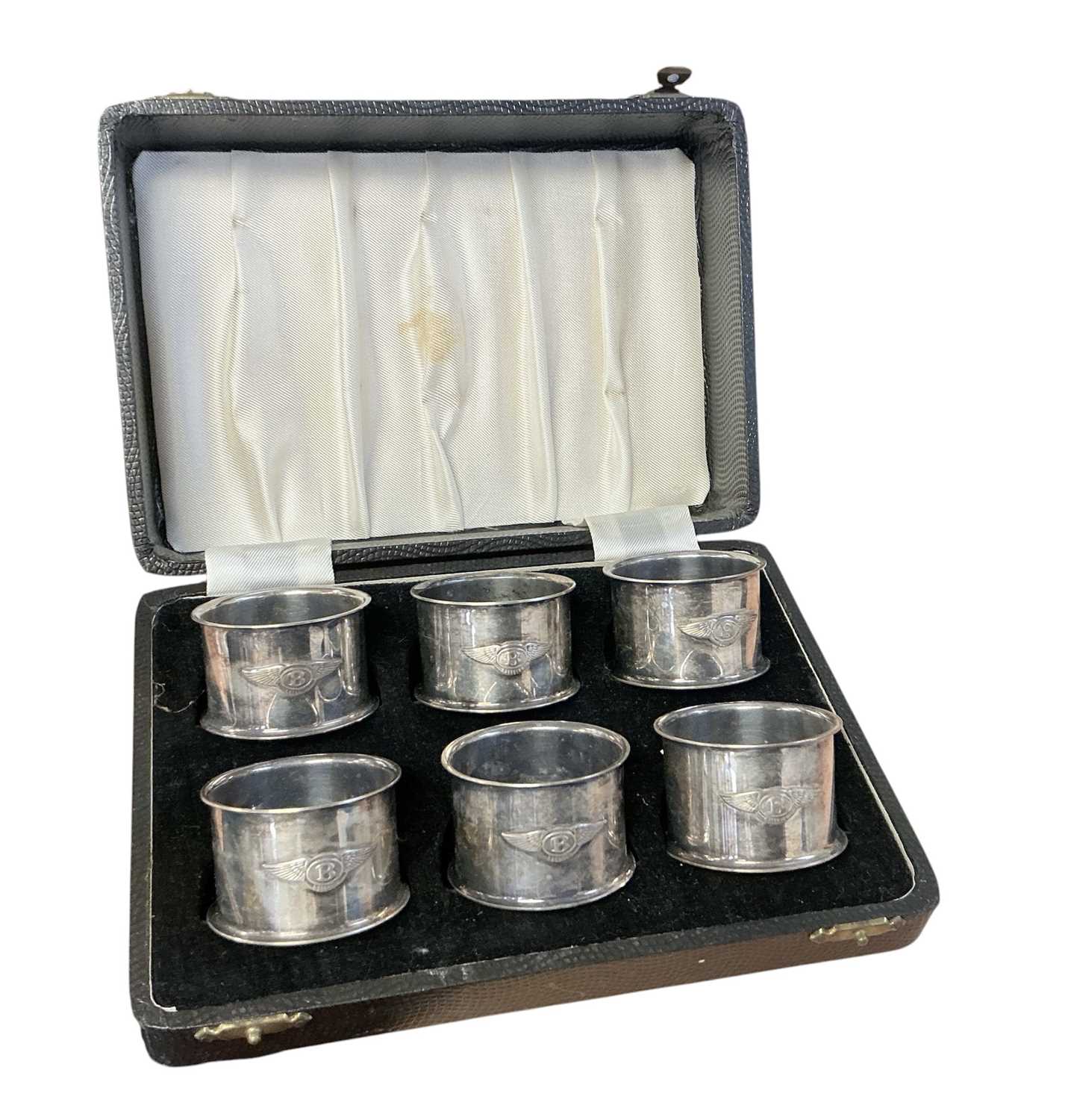Lot 94 - Set of six 1930s silver plated napkin rings, each mounted with a Bentley winged B logo, in a fitted case